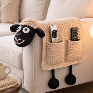 May include: A beige crochet sheep-shaped sofa arm organizer with two pockets for remote controls. The sheep has a black face, ears, and legs. The organizer is attached to a beige sofa.