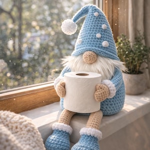 Puede incluir: Una muñeca de gnomo de ganchillo hecha a mano con un gorro y botas de color azul claro, sosteniendo un rollo de papel higiénico. El gnomo tiene barba blanca, nariz y manos color beige, y pompones blancos en el gorro. Está sentado en un alféizar.