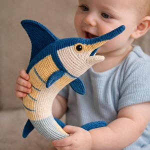 May include: A blue, yellow, and cream-colored crochet marlin fish toy. The toy has a long, pointed snout, a dorsal fin, and a curved tail. The toy is being held by a baby wearing a blue shirt.