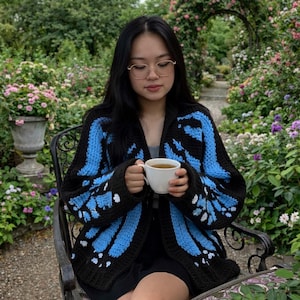 Könnte beinhalten: Ein schwarzer Häkelcardigan mit blauen Schmetterlingsflügelmustern. Der Cardigan hat weiße Details und wird von einer Person getragen, die eine weiße Tasse hält. Der Hintergrund zeigt einen Garten mit Blumen und Grün.