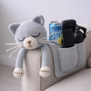 May include: A light blue crocheted cat-shaped sofa arm organizer with two pockets. The cat has closed eyes, a beige nose, and white whiskers. A can of Sparkling water, a phone, and headphones are in the pockets.