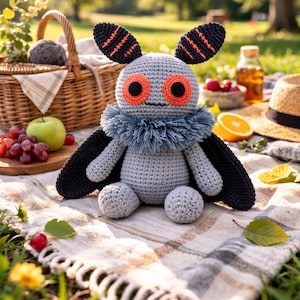 Könnte beinhalten: Handgemachtes, graues Häkel-Motten-Plüschtier mit schwarzen Flügeln und orangefarbenen Akzenten. Das Plüschtier hat große orangefarbene Augen und einen blauen Fransenkragen. Es sitzt auf einer Picknickdecke mit einem Picknickkorb und Obst im Hintergrund.