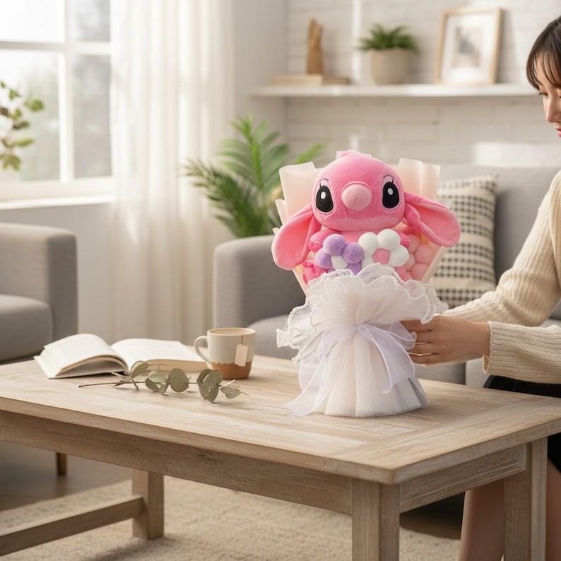 Puede incluir: Un ramo de peluche rosa con un personaje de Stitch con orejas grandes y ojos negros. El ramo incluye detalles florales blancos y morados, envuelto en papel rosa y tul blanco. El ramo est&aacute; sobre una mesa de madera.