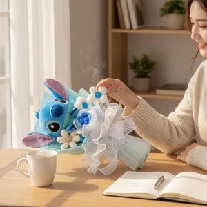 Peut inclure: Un bouquet avec un personnage Stitch en peluche bleu, des fleurs d&eacute;coratives blanches et bleues, et un ruban blanc. Le bouquet est envelopp&eacute; dans du papier bleu clair. Une tasse blanche et un carnet sont sur la table en bois.