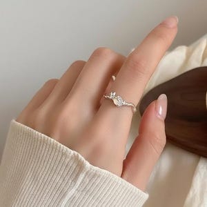 May include: A silver ring with a floral design, featuring a small, round gemstone. The ring is worn on a finger, showcasing its delicate details and elegant style. The ring's design is simple yet eye-catching.