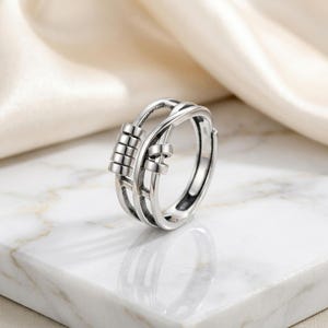 May include: A silver-colored fidget ring with a unique design. The ring features a series of small, movable beads and a woven pattern. The ring is displayed on a white marble surface, with a soft, off-white fabric in the background.