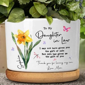May include: White ceramic planter with a watercolor daffodil design and the text "To My Daughter in Law." The planter includes a wooden base and is decorated with butterflies and dragonflies. The text includes the name "Sophia" and a message from "Love, Mom."
