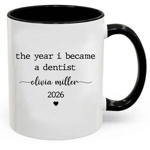 May include: White ceramic mug with a black interior and handle. The mug features the text "the year i became a dentist" with the name "olivia miller" and the year "2026" below a heart symbol.