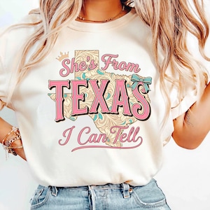 Puede incluir: Camiseta color crema con el texto "She's From TEXAS I Can Tell" en rosa y turquesa. El diseño presenta un mapa de Texas con patrones florales y una corona.