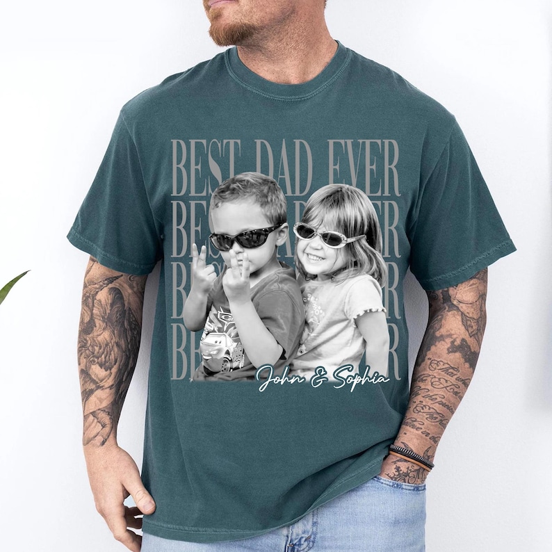 May include: Teal t-shirt with a black and white photo of two children wearing sunglasses. The shirt has the text "BEST DAD EVER" in a repeating pattern and the names "John & Sophia" in a script font.