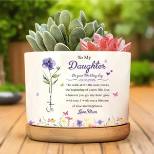 Puede incluir: Una maceta de cerámica blanca con base de madera, llena de suculentas. La maceta está decorada con ilustraciones florales, mariposas y el texto "To My Daughter" y un mensaje de boda. Un regalo reflexivo.