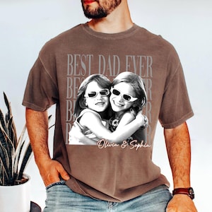 May include: Brown t-shirt with a black and white photo of two girls wearing sunglasses, hugging each other. The text "BEST DAD EVER" is printed in a faded gray font above the photo. The names "Olivia & Sophia" are printed below the photo.
