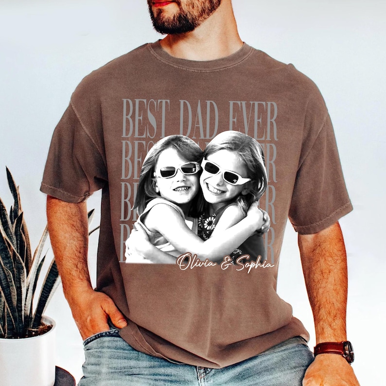May include: Brown t-shirt with a black and white photo of two girls wearing sunglasses, hugging each other. The text "BEST DAD EVER" is printed in a faded gray font above the photo. The names "Olivia & Sophia" are printed below the photo.
