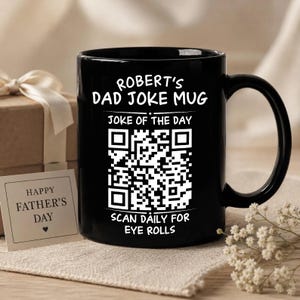 Könnte beinhalten: Ein schwarzer Keramikbecher mit weißem Text, der ROBERT'S DAD JOKE MUG, JOKE OF THE DAY und SCAN DAILY FOR EYE ROLLS lautet, zentriert um einen großen weißen QR-Code. Der Becher hat einen geschwungenen Henkel und eine glatte, glänzende Oberfläche.