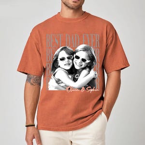 May include: Burnt orange t-shirt with a graphic of two girls wearing sunglasses, hugging. The text "BEST DAD EVER" is printed above the image, with the names "Olivia & Sophia" below. The shirt is a relaxed fit.
