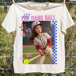 May include: Cream-colored t-shirt featuring a baseball-themed graphic. The graphic includes the text "Hit the Dang Ball" above an illustration of a baseball player doll. Pink and red stars and a blue and white checkered border complete the design.