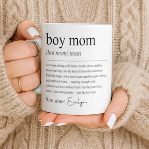 May include: White ceramic mug with the text "boy mom" and a definition. The mug is held by a person wearing a beige sweater. The definition describes a boy mom as a woman raising boys.