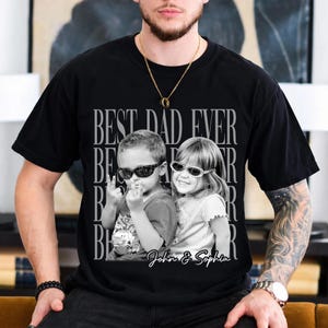 May include: Black t-shirt with a grayscale image of two children wearing sunglasses. The text "BEST DAD EVER" is printed above the image, with the names "John & Sophia" below. The shirt is worn by a person.