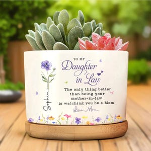 Könnte beinhalten: Ein weißer Keramik-Pflanztopf mit Holzsockel, gefüllt mit grünen und roten Sukkulenten. Der Topf ist mit floralen Designs und dem Text "TO MY Daughter in Law" verziert. Der Text enthält auch "Sophia" und "Love, Mom".