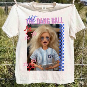 May include: Cream-colored t-shirt with the text "Hit the Dang Ball" in pink and blue. The shirt features a doll wearing a baseball uniform with the number 13. Pink and red stars are also on the shirt.