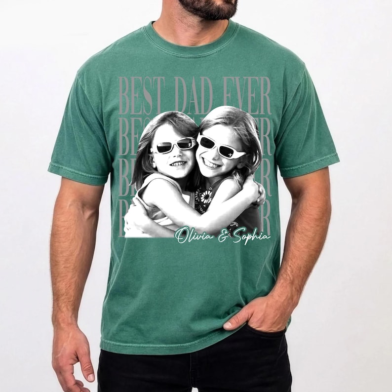 May include: Green t-shirt with the text "BEST DAD EVER" in a faded gray font. The shirt features a black and white image of two girls wearing sunglasses, hugging each other. The names "Olivia & Sophia" are written below the image.