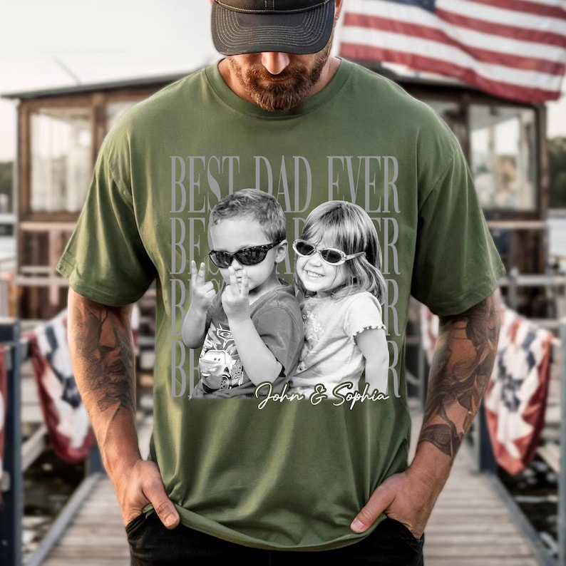May include: Olive green t-shirt with a black and white image of two children wearing sunglasses. The text "BEST DAD EVER" is printed above the image, with the names "John & Sophia" below.