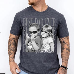 May include: Dark gray t-shirt featuring a black and white photo of two children wearing sunglasses. The text "BEST DAD EVER" is printed above the photo, with the names "John & Sophia" below. The shirt is a crew neck.