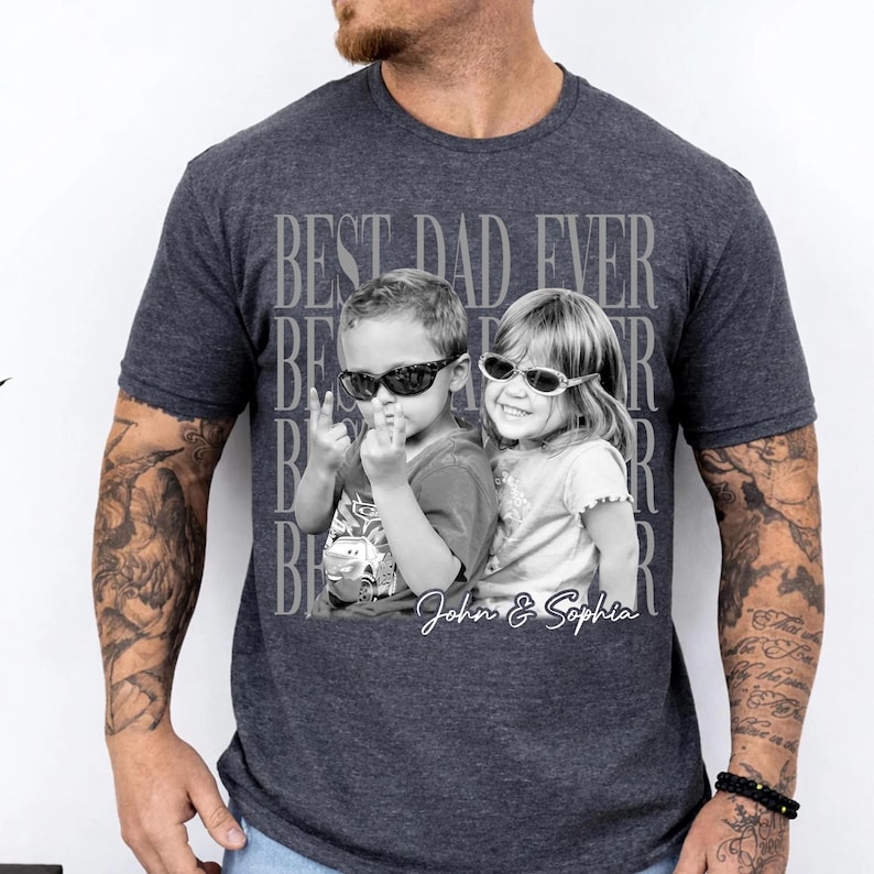 May include: Dark gray t-shirt featuring a black and white photo of two children wearing sunglasses. The text "BEST DAD EVER" is printed above the photo, with the names "John & Sophia" below. The shirt is a crew neck.