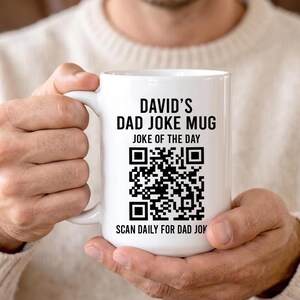Op de afbeelding: Een witte keramische mok met zwarte bedrukte tekst die DAVID'S DAD JOKE MUG, JOKE OF THE DAY en SCAN DAILY FOR DAD JOKES leest. Een grote zwart-witte QR-code is gecentreerd op de voorkant van de mok tussen de tekstregels.