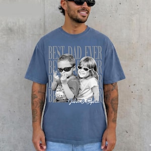 May include: Blue t-shirt with a graphic that says "BEST DAD EVER" in a repeating pattern. The graphic includes a black and white photo of two children wearing sunglasses. The names "John & Sophia" are written below the photo.