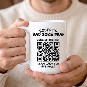 May include: A white ceramic mug featuring black text that reads ROBERT'S DAD JOKE MUG, JOKE OF THE DAY, and SCAN DAILY FOR EYE ROLLS. A large black and white QR code is printed in the centre of the mug between the text lines.