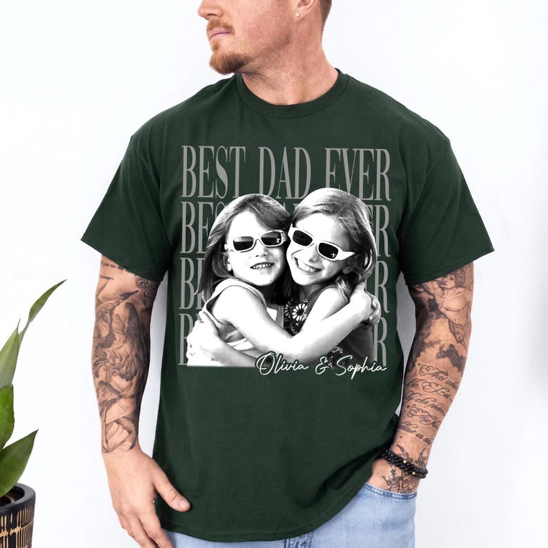 May include: Dark green t-shirt featuring a black and white photo of two girls wearing sunglasses, hugging each other. The text "BEST DAD EVER" is printed above the photo, with the names "Olivia & Sophia" below.