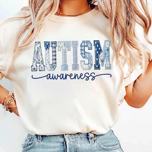 Puede incluir: Camiseta color crema con las palabras "AUTISM awareness" en azul. La palabra "AUTISM" tiene un diseño de patchwork con cuadros azules, flores y lunares. La palabra "awareness" está escrita en cursiva.