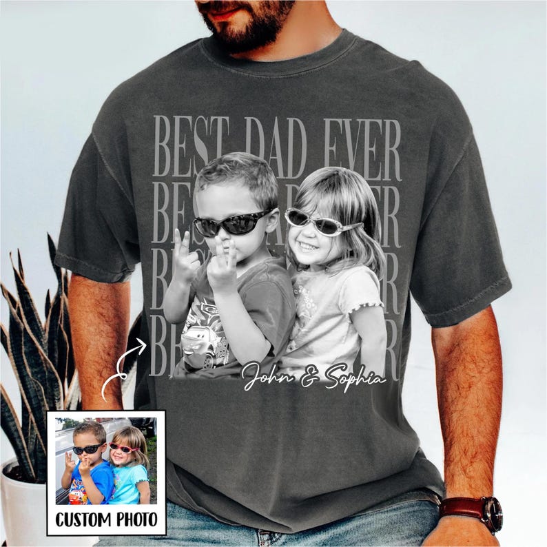 May include: Dark gray t-shirt with a black and white image of two children wearing sunglasses. The shirt has the text "BEST DAD EVER" and the names "John & Sophia" printed on it. A small photo is in the lower left corner.
