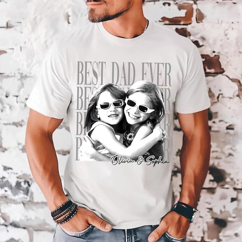 May include: White t-shirt with a black and white graphic of two smiling children wearing sunglasses, hugging each other. The text "BEST DAD EVER" is printed above the image, with the names "Olivia & Sophia" below.