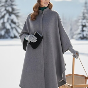 Może przedstawiać: Osoba w szarej pelerynie, czapce i rękawiczkach stoi w zaśnieżonym krajobrazie, trzymając czarną kopertówkę i ciągnąc drewniane sanki. Peleryna ma zapięcie na guziki i zaokrąglony dół. W tle ośnieżone drzewa i góry.