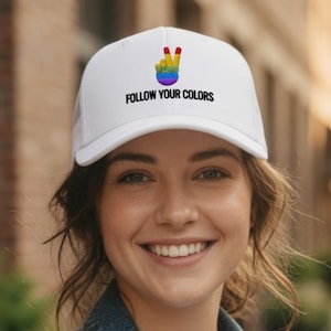 Könnte beinhalten: Weiße Trucker-Cap mit einem Regenbogen-Friedenszeichen und dem Text "FOLLOW YOUR COLORS". Die Kappe hat einen Mesh-Rücken und einen gebogenen Schirm. Das Friedenszeichen ist in den Farben des Regenbogens. Der Text ist schwarz.