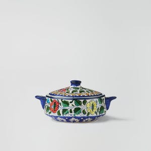 May include: A ceramic serving dish with a lid, featuring a vibrant floral pattern in red, yellow, and green on a white background. The dish has blue handles and a blue rim, perfect for serving food.