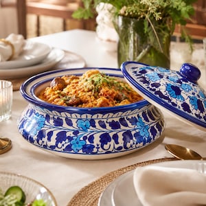 May include: A round ceramic serving bowl with a matching lid, featuring a white base decorated with hand-painted blue floral and vine patterns. The bowl is filled with a cooked grain dish topped with green herbs, sitting on a table setting.