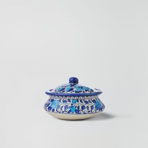 Puede incluir: Un cuenco de cerámica con tapa, con un diseño floral azul sobre fondo blanco. El cuenco tiene forma redondeada con tapa a juego y un pequeño pomo. El diseño incluye varios tonos de azul, creando una pieza decorativa y funcional.