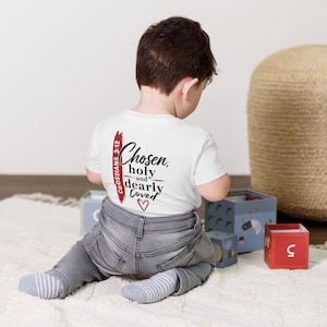Op de afbeelding: Een wit T-shirt met korte mouwen voorzien van de zwarte tekst Chosen, holy and dearly loved, een rood hart en een verticale rode penseelstreek met de tekst Colossians 3:12. Het shirt wordt gedragen met een grijze spijkerbroek en gestreepte sokken.