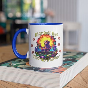 Könnte beinhalten: Weiße Keramiktasse mit blauem Griff und Innenraum. Die Tasse zeigt eine farbenfrohe Illustration eines lila Affen, der ein Getränk hält, mit dem Text "Monkey See Monkey Brew" in einer skurrilen Schriftart. Die Tasse steht auf einem Stapel Bücher.