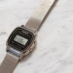 Op de afbeelding: Een zilverkleurig digitaal polshorloge met een rechthoekige wijzerplaat en een fijne metalen mesh-band. De zwarte wijzerplaat toont de merknaam SKMEI, de tijd 11:06 en labels voor alarm, timer, mode, start, stop en count down timer functies.