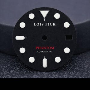 Può includere: Un quadrante di orologio nero rotondo con indici circolari bianchi, un indice triangolare a ore dodici e indici rettangolari a ore sei e nove. Il testo LOIS PICK è stampato in bianco, con PHANTOM AUTOMATIC in rosso e bianco.
