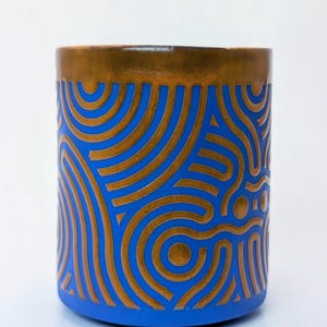 Könnte beinhalten: Ein zylindrischer Keramikbehälter mit blauem Grund und einem bronzefarbenen Rand. Die blaue Oberfläche ist mit einem sich wiederholenden Muster aus bronzefarbenen geschwungenen Linien und Kreisen verziert. Der Behälter ist wahrscheinlich ein Dekorationsartikel oder eine kleine Vase.
