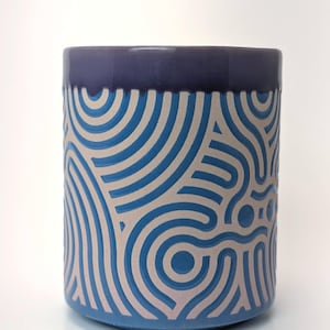 May include: A cylindrical ceramic container with a blue base and a dark blue rim. The body features a repeating pattern of light blue and off-white curved lines. The design creates a sense of movement and visual interest. The container is suitable for holding items.
