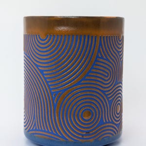 Könnte beinhalten: Zylindrische Keramiktasse mit blauem Boden und braunem Rand. Die Tasse hat ein sich wiederholendes Muster aus kupferfarbenen konzentrischen Kreisen auf blauem Hintergrund. Das Design erzeugt einen wellenartigen Effekt.
