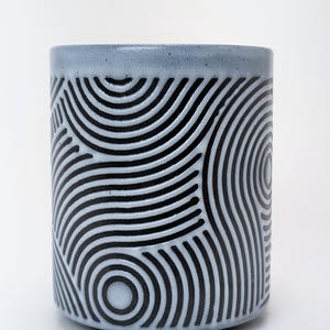 Könnte beinhalten: Ein zylindrischer Keramikbehälter mit hellblauem Rand und einem Muster aus schwarzen und weißen konzentrischen Kreisen. Das Design bedeckt den gesamten Körper des Behälters. Der Behälter wird wahrscheinlich zur Aufbewahrung oder Präsentation verwendet. Höhe ca. 10 cm.