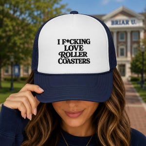 May include: A navy blue and white trucker hat featuring a foam front panel with the black printed text I F*CKING LOVE ROLLER COASTERS. The cap has a solid navy blue brim and a mesh back panel, designed with a structured crown and an adjustable snapback.