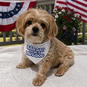 May include: A small, light brown, long-haired dog wearing a white triangular bandana around its neck. The bandana features the blue text 'BAD DAY TO BE A HOT DOG' printed in a bold, rounded font. The dog has floppy ears and dark eyes.